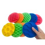 Kit de 10 discos sensoriais táteis coloridos sobre um fundo claro. Discos têm texturas variadas e estão organizados em duas fileiras, junto a um tapa-olho preto.