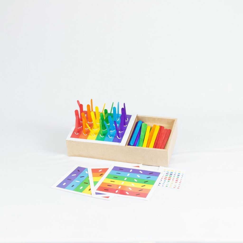 Kit Caixa de Palitos Coloridos da Materiais Para Brincar, mostrando uma caixa de MDF com alguns palitos já inseridos nos orifícios. Uma cartela com um desafio de cores está visível ao lado.
