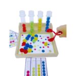 O jogo Sequência de Pompons da Materiais Para Brincar, mostrando a caixa de madeira com os 4 tubos de ensaio. Uma mão de criança está usando a pinça bola para colocar um pompom colorido em um dos tubos, seguindo o padrão de uma das fichas modelo.
