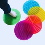 Kit Tapete Sensorial da Materiais Para Brincar, mostrando os 5 tapetes coloridos e texturizados (com pontinhos, ondas, etc.) dispostos lado a lado no chão. Uma criança pode estar descalça sobre um dos tapetes.
